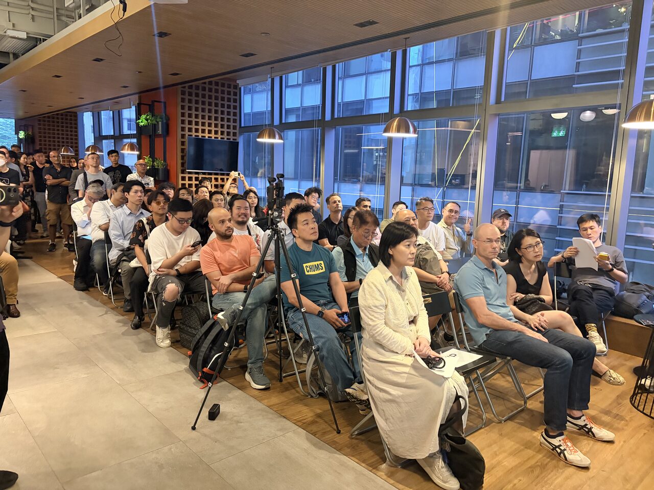 A packed audience seated and watching, city windows behind