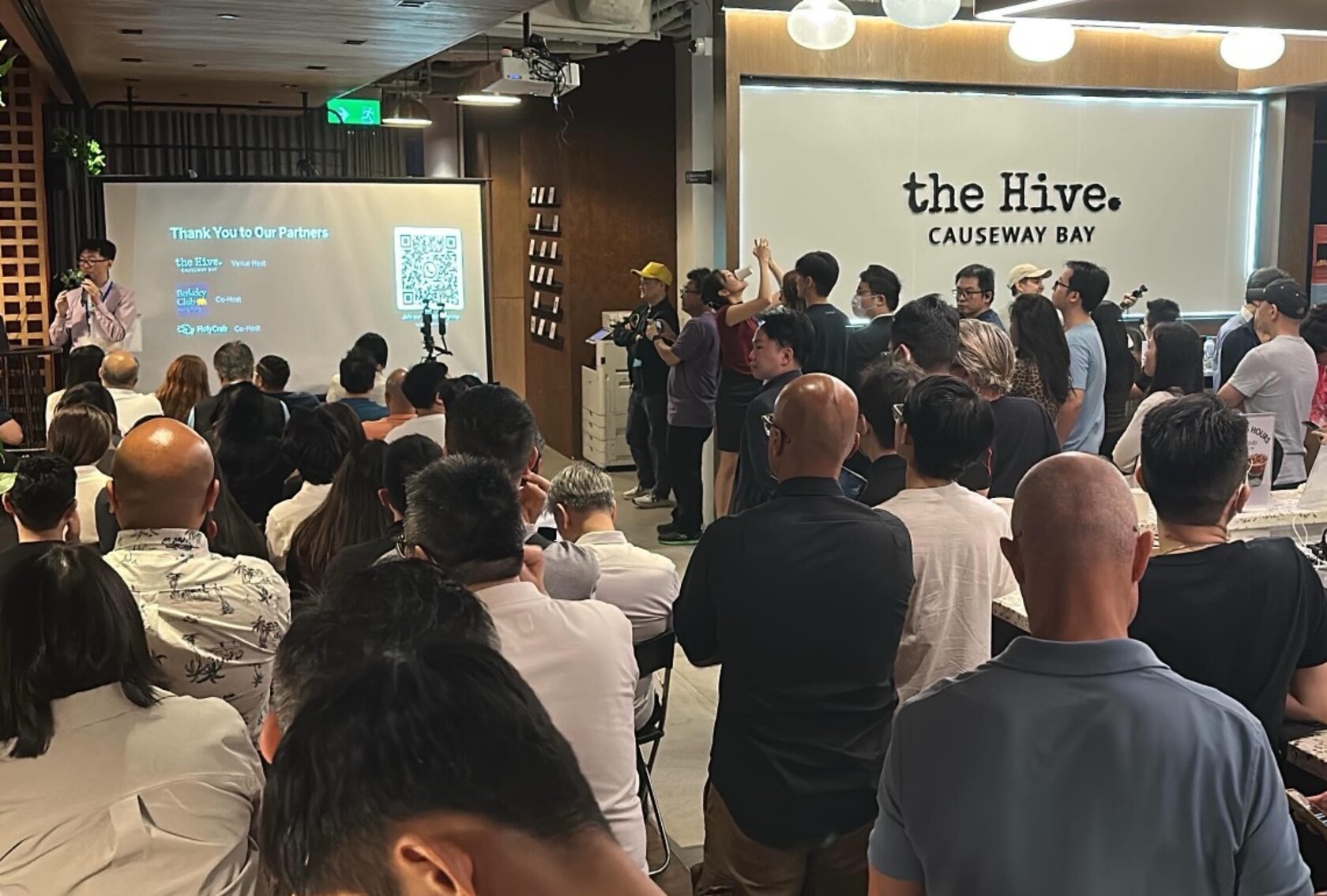 A packed room at The Hive Causeway Bay watching the talks, the venue signage lit up behind the crowd